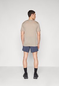 Men’s short-sleeve beige t-shirt, back view, paired with navy shorts, black socks, and dark athletic shoes, set against a neutral background.