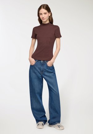 Top corto aderente marrone a maniche corte abbinato a jeans blu larghi. La modella è in piedi con le mani in tasca, indossando sneakers grigie con dettagli colorati.