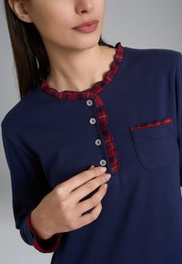 Maglietta a maniche lunghe di colore blu navy con rifiniture a quadretti rossi, scollatura ondulata, sei bottoni frontali e una tasca con dettaglio a quadretti coordinato.