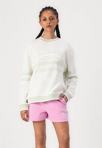 Felpa bianca a maglia waffle con strisce verdi e logo ricamato, abbinata a pantaloni corti rosa con stampa logo, che mettono in mostra un design casual.