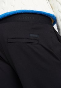 Svarta byxor med en slät yta, med en bakficka som har en liten svart Calvin Klein-etikett. Midjan har präglad logotyp.