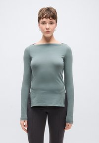 Hosszú ujjú felső puha, visszafogott zöld anyagból, csónaknyakú és testhez simuló szabású, sötét, karcsúsított nadrággal párosítva.