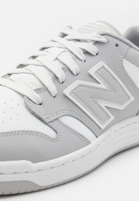 Hvide og grå sneakers med læder- og tekstilmaterialer, synlige syninger, fremtrædende logo, teksturerede såler og traditionel snørebåndslukning.