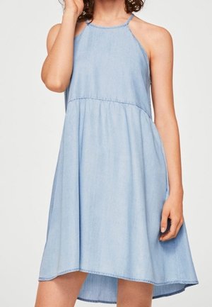 Mujer con vestido sin mangas de mezclilla azul claro con escote halter y falda acampanada, de pie contra un fondo liso.