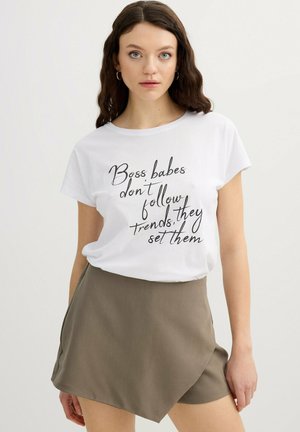 Mujer con cabello castaño lleva una camiseta blanca con la frase "Las jefas no siguen las tendencias, las crean" y una falda marrón asimétrica.