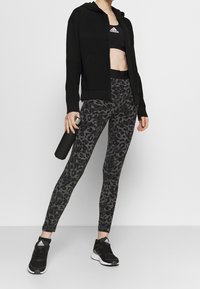 Felpa nera con zip e parte superiore testurizzata, reggiseno sportivo nero, leggings grigi con stampa leopardata e scarpe da ginnastica nere, mentre tiene una bottiglia d'acqua.
