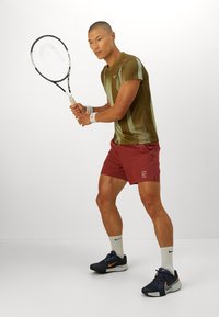 Idrettsutøver i en stripete brun skjorte og røde shorts klar til å slå en tennisball med et racket mot en ensfarget hvit bakgrunn.