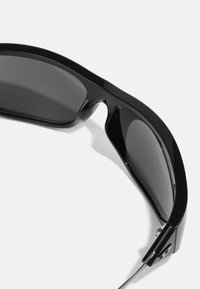 Sorte solbriller med et str�ømlinet, vinklet design. Har mørke, reflekterende linser og teksturerede sorte rammer med sølvdetaljer på hængslerne.