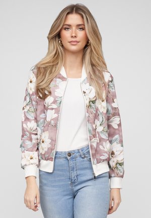 Kobieta ubrana w jasnoróżową kwiecistą kurtkę bomberkę na białej bluzce i jasnoniebieskich dżinsach, stojąca na tle jednolitego szarego tła.