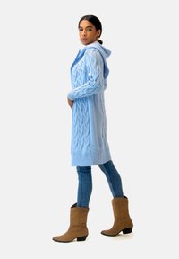 Ljusblå stickad huvtröja med kabelstickning, långa ärmar och ribbade muddar, som bärs tillsammans med figursydda blå jeans och bruna stövlar.