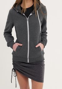 Grey zip-up hoodie made from soft fabric, featuring a front zipper, drawstrings, and two front pockets. Paired with a black, ruched mini skirt.