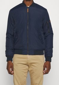 Giubbotto bomber blu navy con zip, polsini a coste, tasche laterali e tasca sul braccio con dettaglio in pelle. Tessuto dalla consistenza liscia.