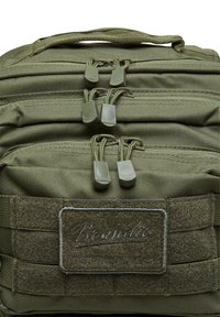 Olivgrön taktisk ryggsäck med justerbara remmar, två fickor med dragkedja med gummihandtag och en strukturerad Velcro-lapp för anpassning.