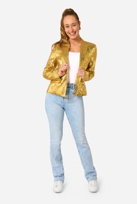 Zelta spīguļota žakete ar pieguļošu dizainu, apvienota ar gaiši ziliem džinsiem un baltiem sporta apaviem. Modelis stāv ar rotaļīgu izteiksmi.