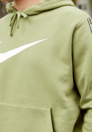 Osoba nosząca jasnozieloną bluzę z kapturem z białym logo Nike Swoosh i widocznymi sznurkami.