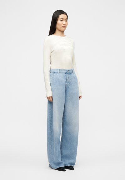 Γυναίκα που στέκεται, φορώντας ένα μακρυμάνικο εκρού τοπ με ανάγλυφη ύφανση, ανοιχτό μπλε τζιν παντελόνι με φαρδιά πόδια και μαύρα μυτερά παπούτσια σε λευκό φόντο.