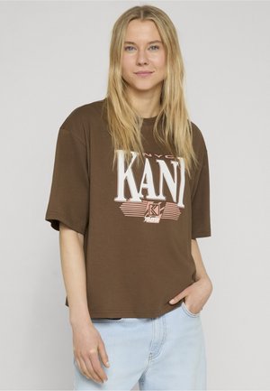 Blondynka z luźno rozpuszczonymi włosami, ubrana w brązowy, oversize'owy t-shirt KANI i jasnoniebieskie dżinsy, stojąca z jedną ręką w kieszeni.