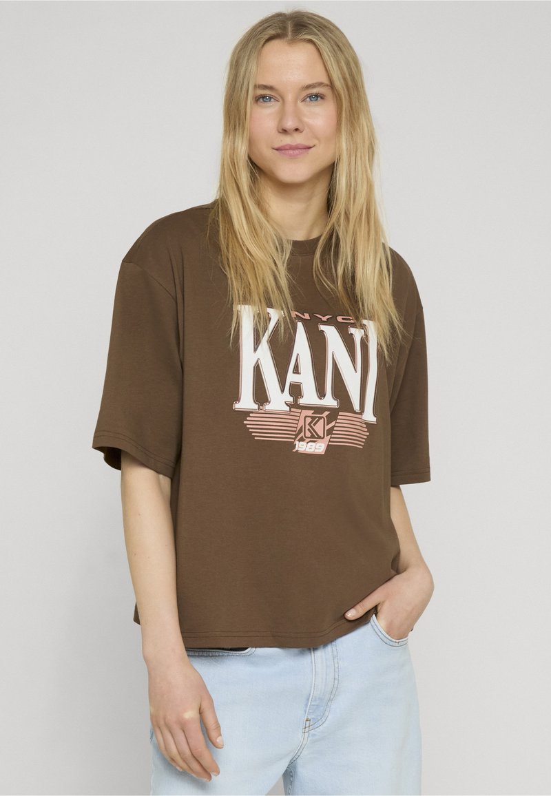 Blonde vrouw met los haar, die een bruin oversized KANI T-shirt en lichtblauwe spijkerbroek draagt, staande met één hand in de zak.