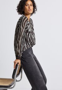 Blusa a strisce zebra nera e grigia trasparente con maniche ampie abbinata a jeans grigio scuro, che tiene in mano una sedia minimalista.
