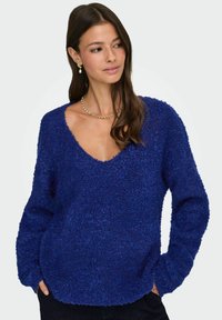 Modra pulover z teksturo in globokim V-izrezom, dolgimi rokavi in sproščenim krojem, ki prikazuje mehak, puhast material in ukrivljen rob.