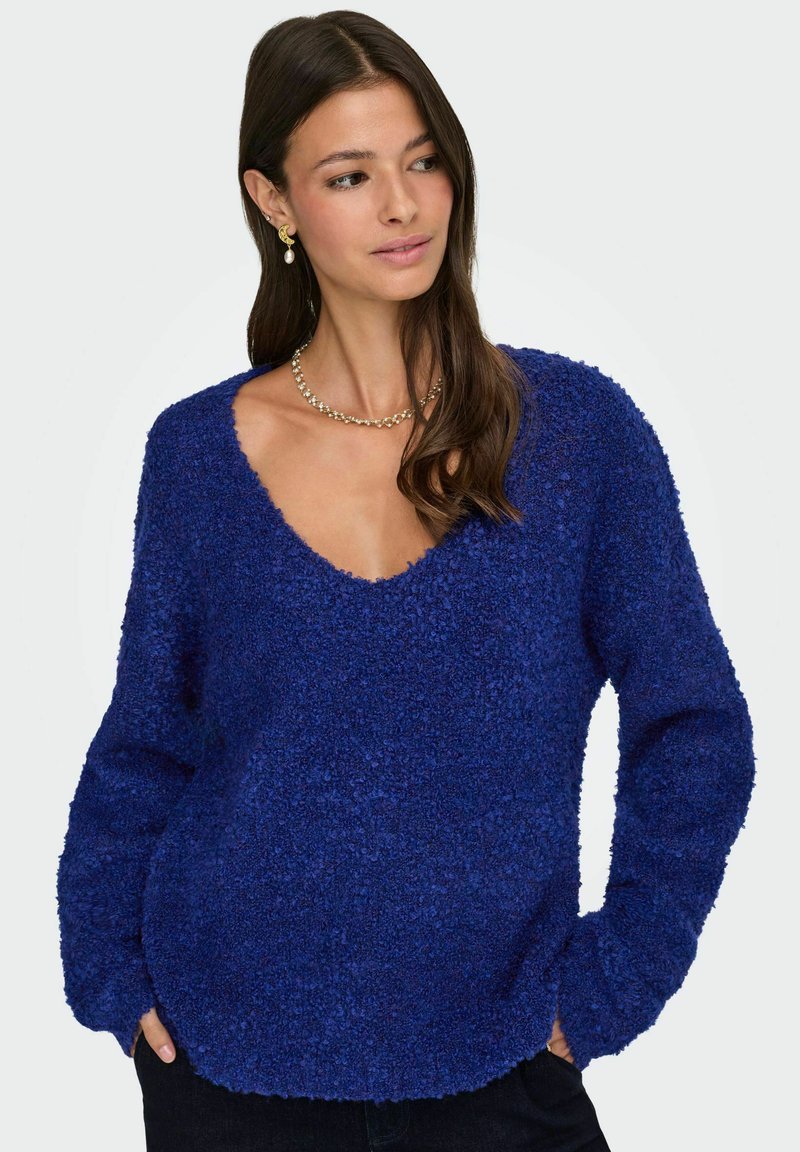 Modra pulover z teksturo in globokim V-izrezom, dolgimi rokavi in sproščenim krojem, ki prikazuje mehak, puhast material in ukrivljen rob.