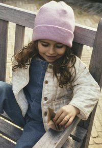 Padded krem jopica z rjavimi gumbi, jeans kombinezoni in roza pletena kapa; otrok drži majhen rjav papirni predmet medtem ko sedi.