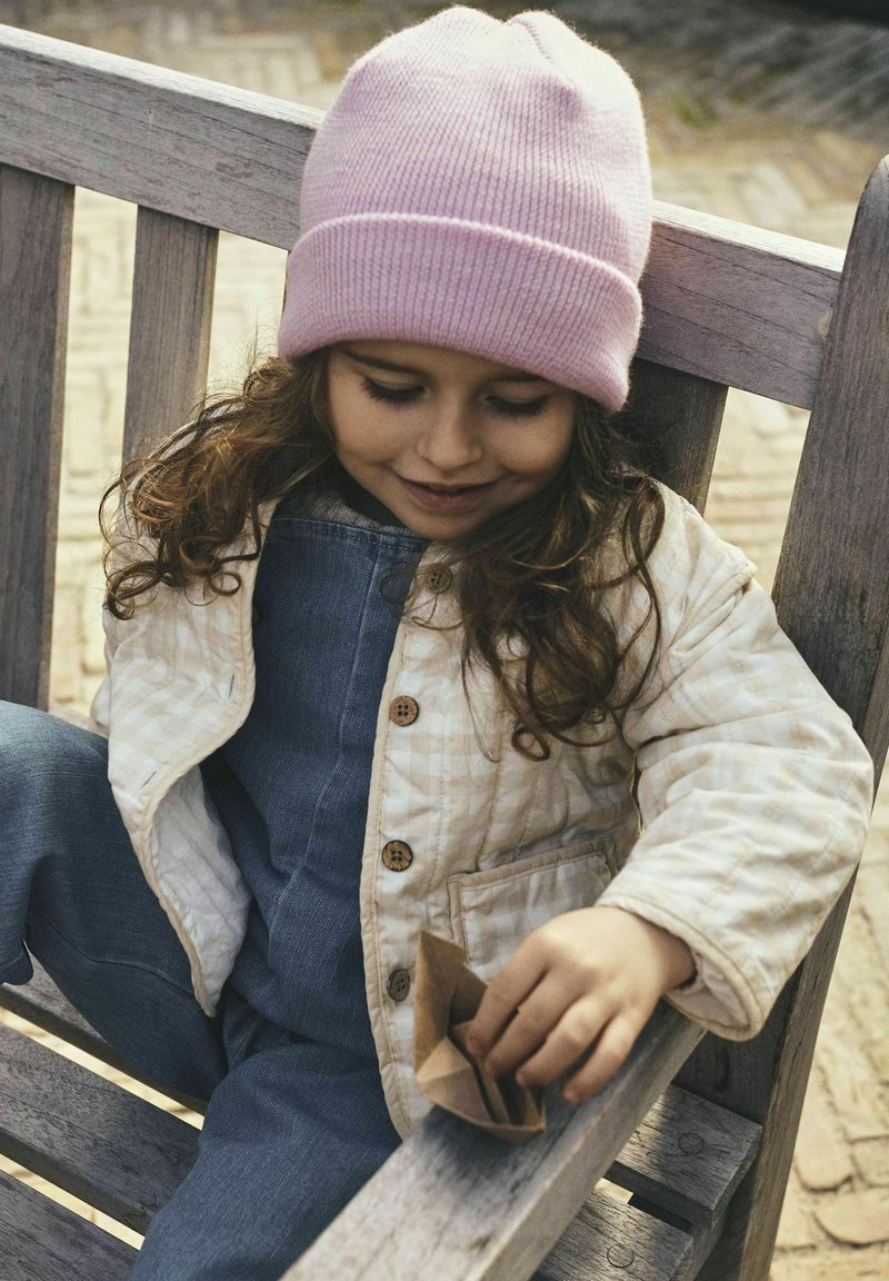 Padded krem jopica z rjavimi gumbi, jeans kombinezoni in roza pletena kapa; otrok drži majhen rjav papirni predmet medtem ko sedi.