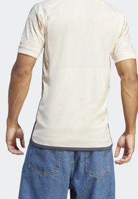 Camisa atlética de un beige claro con mangas cortas, textura sutil y un ribete gris en el dobladillo. Llevada con pantalones cortos de mezclilla azul.