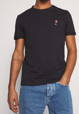 Czarna bawełniana koszulka z małym różowym haftowanym różą na klatce piersiowej, okrągłym dekoltem i podwiniętymi krótkimi rękawami. W połączeniu z niebieskimi dżinsami.