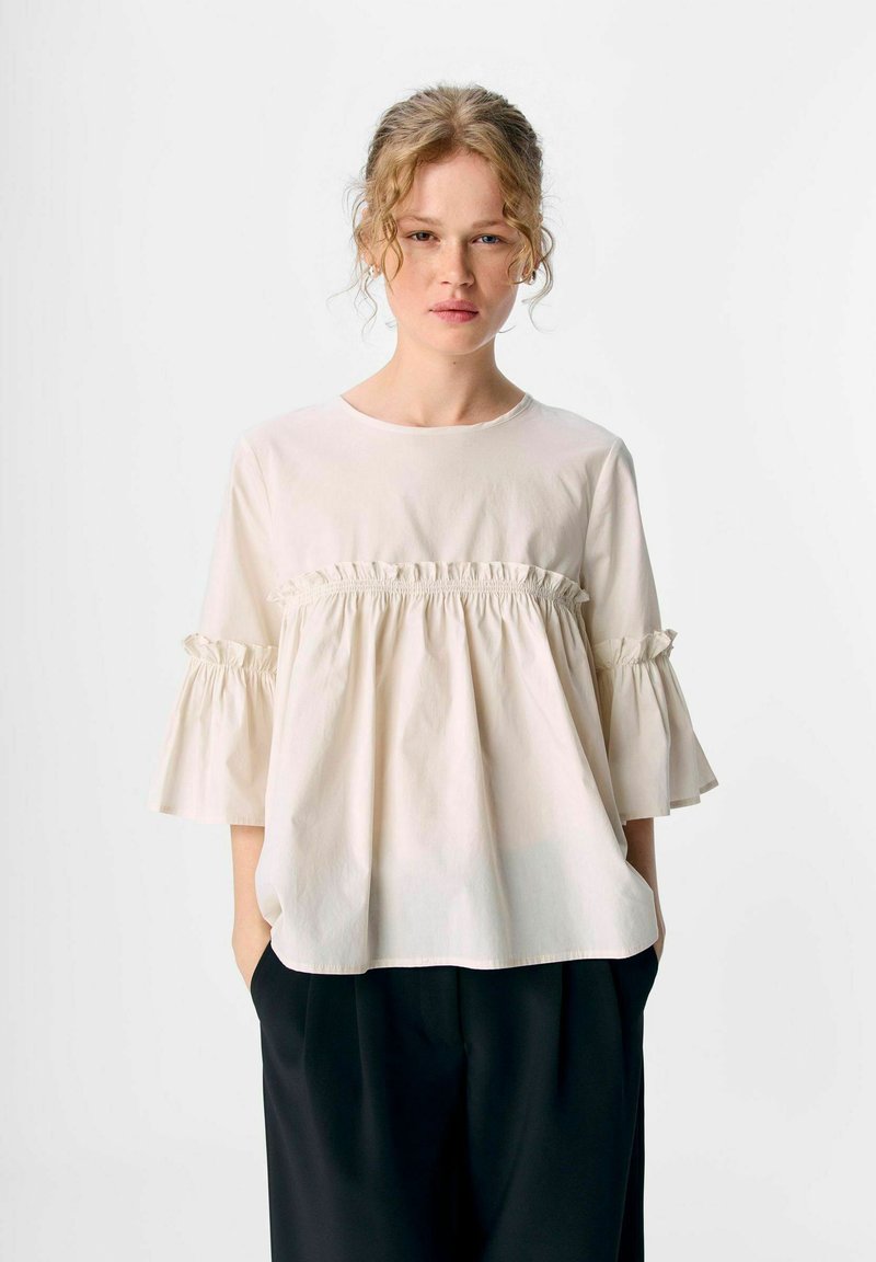 Femme aux cheveux bouclés portant une blouse beige ample avec des coutures froncées et des manches larges, associée à un pantalon noir, se tenant devant un fond uni.