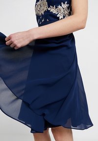 Marinblå chiffongklänning med en åtsittande liv och flödande kjol, prydd med blommig broderi och en vågig fåll.