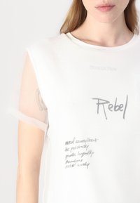 Maglietta bianca con maniche trasparenti, caratterizzata da un collo rotondo e testo stampato in grigio. Dettagli notevoli includono "Rebel" e varie frasi scritte a mano.
