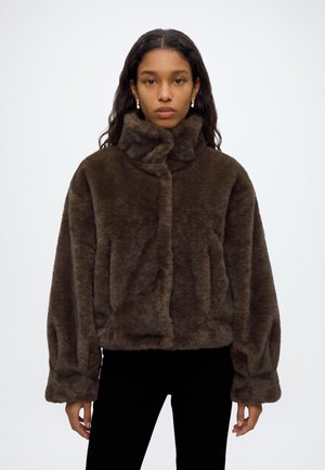Femme aux cheveux foncés portant une veste en fausse fourrure brun foncé à col montant et un pantalon noir sur un fond clair uni.