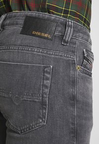 Primer plano del bolsillo trasero y la cinturilla de unos jeans Diesel grises con etiqueta de marca de cuero, combinados con una camisa de cuadros verde y roja metida por dentro.