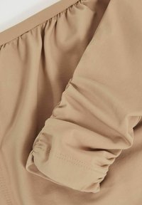 Beige kledingstuk met een geplooid manchet detail, gemaakt van zachte stof. De textuur lijkt zacht met subtiele stiksels langs de randen.