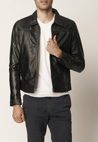 Giacca di pelle nera con texture liscia, chiusura con zip e colletto. Abbinata con una camicia bianca e pantaloni scuri.