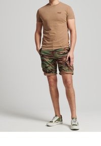 Man som bär en beige kortärmad tröja, kamouflage cargo-shorts och gröna och vita sneakers, står med händerna i fickorna på en grå bakgrund.