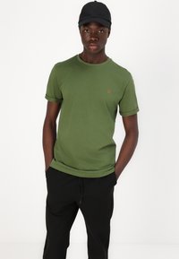 Green cotton t-shirt with rolled short sleeves and small orange logo, paired with black drawstring trousers. Casual fit and simple design.