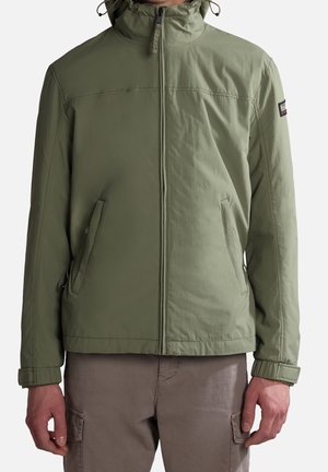 Mann trägt hellgrüne Zip-up Jacke mit Kapuze und braune Cargohosen, gezeigt vom Hals bis zur Hüfte vor einfachem Hintergrund.