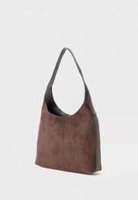 Sac à épaule en suede marron avec des finitions et une sangle en cuir marron foncé, posé debout sur un fond blanc.