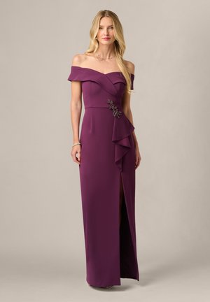 Blonde vrouw draagt een paarse off-shoulder avondjurk met een decoratieve broche en een voorste split, staand tegen een effen achtergrond.
