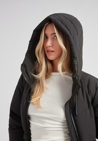 Ung kvinde med langt blond hår iført en sort polstret jakke med hætte over en hvid tætsiddende top, der ser lidt til siden.