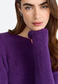 Purple knitted sweater with a crew neck and long, ribbed cuffs. Soft texture with a slightly fuzzy finish. Model's hand rests on cheek.