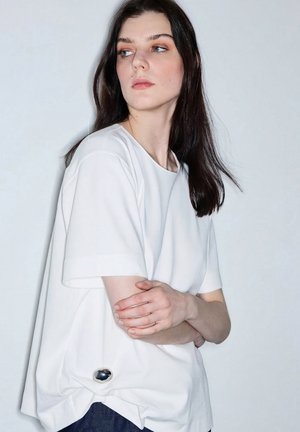 Joven con el pelo largo y oscuro, vestida con una camiseta blanca holgada, mirando de lado con los brazos cruzados, sobre un fondo claro y liso.