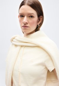 Νεαρή γυναίκα που φοράει μια εκρού κοντομάνικη μπλούζα με κουμπιά και ένα μάλλινο κασκόλ που καλύπτει τους ώμους της, μπροστά σε απλό φόντο.