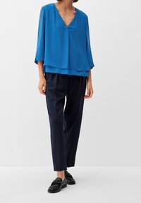 Personne portant un chemisier bleu ample à col en V, un pantalon tailleur foncé et des mocassins en cuir noir, debout devant un fond blanc uni.