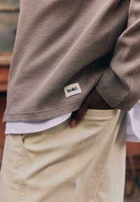 Personne portant un pantalon beige et une chemise à manches longues taupe texturée avec une petite étiquette blanche « solid. » près de l'ourlet, main dans la poche.