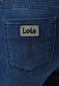 Temno modre kavbojke iz jeansa z zadnjim žepom, na katerem je svetlo modra kvadratna nalepka z napisom "Lois" v črnem pisavnem slogu.