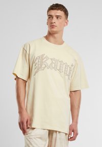 Light yellow cotton t-shirt with short sleeves and a loose fit. Features a textured graphic design in silver reading "Kani."