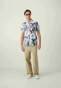 Camisa de botão de manga curta em estampado floral cinza e branco, combinada com calças bege e ténis brancos, usada por um modelo em pé.